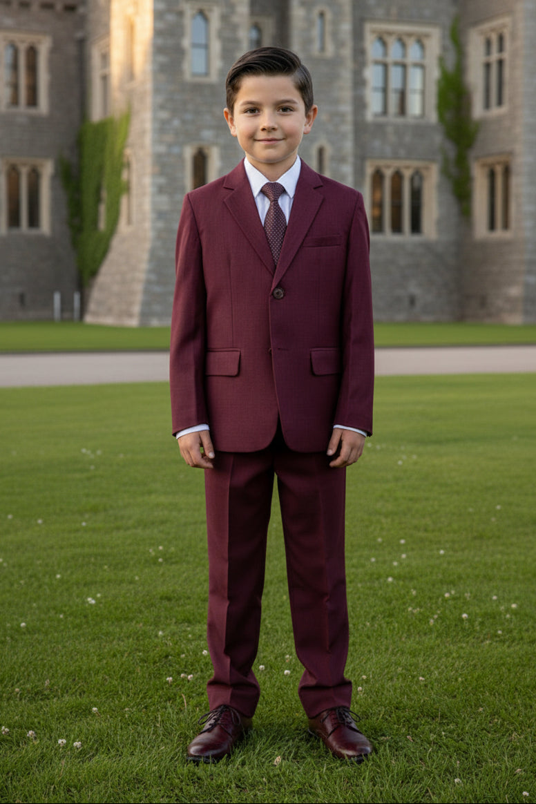 A boys  single-breasted blazer in burgundy with a subtle textured weave, hanging on a black hanger against a plain beige wall. The blazer is styled with a white dress shirt and a patterned burgundy tie.